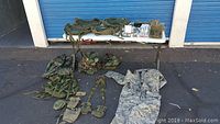 Full view of military lot items on table in front of blue garage doors including jacket, cargo bags, and pouches.