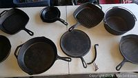 Top view of seven cast iron pans including two square grill pans, three round skillets, one Dutch oven with bail handle, and detached handle, showing wear and age patina.