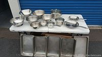 Wide shot of stainless steel kitchenware including mixing bowls, colanders, saucepan, and baking trays arranged on a white table.