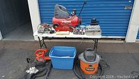 Wide shot of table with Husky air compressor, various tools, Ridgid wet/dry vacuum, and blue plastic bin of hand tools