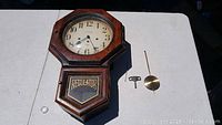 Front view of the vintage wooden wall regulator clock with octagonal face, glass cover, and 'REGULATOR' in gold on lower panel. Includes winding key and pendulum placed on the surface.