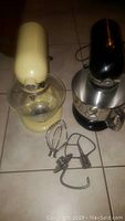 Photo showing both KitchenAid mixers (cream retro K45 and black Artisan KSM150PSOB) side by side on floor with glass and stainless steel bowls and attachments laid out.
