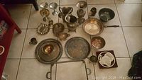 Wide shot showing full lot of 22 tarnished silverplated pieces including trays, cups, goblets, serving dishes and decorative items arranged on tiled floor.