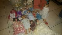 Top view of 18 assorted dolls on tiled floor showing varied clothing and sizes