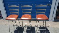 Photo of three iron back bar stools with brown seats lined up outdoors on pavement.