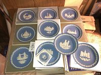 Photo showing all 11 Wedgwood Christmas plates in boxes laid out on floor, each approximately 8 inches diameter. Plates feature various white relief landmark designs on blue backgrounds with white laurel wreath borders.