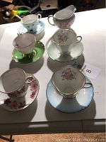 Photo of five porcelain teacups each with matching saucer and one creamer pitcher on a white surface, showing floral designs and gold trims.