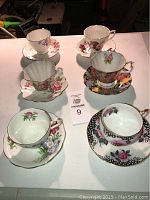 Photo of 6 assorted teacups made in England, each paired with matching saucers, displaying various floral patterns and gold trims