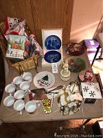 Full view of the Christmas lot items showing dinnerware cups, plate, blue collector plates, porcelain figurines, and Christmas textiles.
