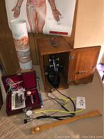 Photo showing the microscope in wooden case, Acupunctoscope device with probes in red case, two stethoscopes with black and yellow tubing, wooden back scratcher laid on table, and a rolled human anatomy poster in the background.