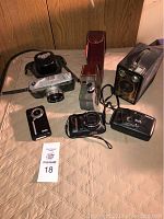 Six assorted vintage cameras arranged on beige surface, various shapes and sizes, some in cases