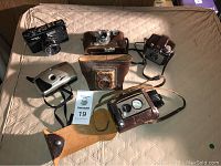 Six assorted cameras displayed on beige fabric surface, including vintage Yashica MG-1, Kodak Brownie, and others, some with cases