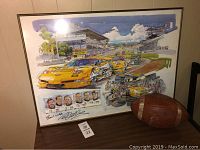 Framed print with yellow race cars on a race track and an old football resting next to it on a wooden surface.