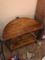 Wooden half circle wall table with carved side supports and two wooden shelves, viewed from an angle showing tabletop and lower shelf.