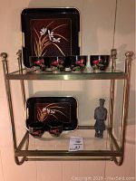 Full view of brass framed glass shelf with two black lacquerware trays and 6 sake tea cups on top shelf, and 3 sake cups and figurine on lower shelf