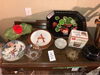 Photo of nine assorted decorative items including vintage tray, Asian collector plate, several glass bowls, Brazilian coin plate, ceramic leaf dish with ladybug, and other small plates and bowls on a dark wooden table.