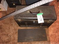 Closed black metal toolbox with brown handle, lock with key, and a metal meter stick lying diagonally across the box.
