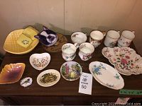 Table display of vintage decorative china and accessories featuring yellow purse and lighter, small decorative plates, and bone china mugs with florals and gold accents