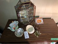 Photo showing small glass curio cabinet with brass framing and various decorative items arranged on a table including a ceramic house teapot, shell bowls, and glass cup.