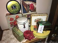 Photo of various retro tins including Lions Club and floral themed tins, a white ceramic canister with latch, and red ceramic piggy bank on a wooden shelf.