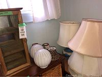 Wooden display cabinet alongside various lampshades and ceramic lamp base, shown near window with natural light