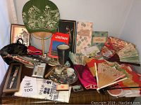 Wide view of lot showing green circular fan, folding fans, ceramic cup, framed art, stationery, and various souvenirs.