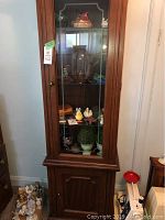 Front view of the tall wooden china cabinet with glass door containing decorative items and lower solid door.