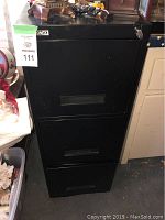 Photo of the full black metal filing cabinet with key in lock on side, showing three drawers and some wear.