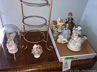 Photo of gold-colored metal plate holder, fruit holder base and several figurines including hummingbird under glass dome, and small flower arrangement figurine
