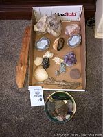 Top view showing assorted geodes, shells, driftwood and large flat polished stone inside a cardboard box with MaxSold measurement scale.