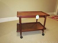 Front side view of teak tea trolley showing two shelves and four black caster wheels on beige carpet.