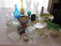 Photo showing assorted glassware and porcelain items including vases, bowls, plates, pitchers, and collectible dishes on white tablecloth.