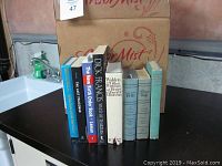 Books standing upright on a table in front of cardboard boxes showing covers of some titles.