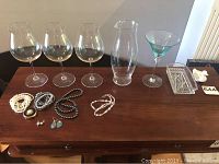 Set of 3 clear wine glasses, 1 clear glass vase, 1 green-stemmed cocktail glass, assorted jewelry (earrings, necklaces, bracelets) on wooden table