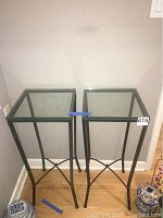 Pair of glass top corner tables with black metal frames and square glass tops placed against a gray wall on hardwood floor.