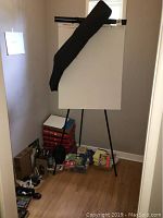 Whiteboard easel set against a corner wall with the black carrying case draped on the top bar. Several boxed and loose office supplies arranged at the base of the easel.