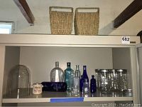 Shelf with 2 rectangular wicker baskets on top, assorted vintage glass bottles and jars below with glass dome and small ceramic container