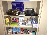 Full shelf view showing roaster pan with rack, blue broiler pan, black pots, and assorted kitchen storage and cleaning supplies on three shelving levels.