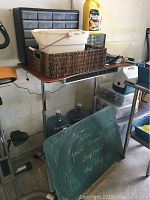 Metal and wood utility table with various items on top including a wicker basket and plastic containers, chalkboard leaning against table