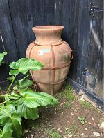 Outdoor terra cotta pot with metal banding and handles, set against wooden fence with green plant nearby.