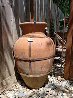 Front view of large terracotta pot with metal bands and ring handles outside on gravel next to wooden fence.