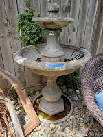 Full view of a large concrete garden fountain with two basins and an electric pump cord coiled in the base basin. Fountain is situated outdoors on stones with a wooden fence background.