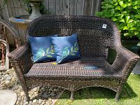 Front view of dark brown woven wicker settee with two blue pillows with bird designs on it, placed outdoors with pebbles under and wooden fence background.