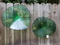 Two green decorative glass yard art pieces hung on a wood fence, showing their translucent green surface and wavy organic edges.