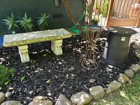 Concrete bench with carved legs beside two different plant stands in a garden bed with mulch and stone border.