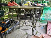 Full view of the outdoor table showing glass surface and metal legs with decorative curved metalwork.