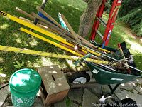 Full view of wheelbarrow filled with assorted garden tools including rakes, shovels, hoes, and poles. Green bucket and wooden box nearby.