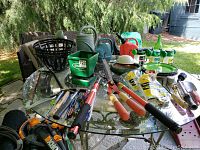 Overview of various garden tools, seed spreader, watering cans, hanging baskets, and hoses laid out on glass table
