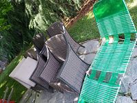 Photo showing four matching woven outdoor chairs with brown metal frames, three beige cushions, and one green plastic lounge chair on stone and grass surface.