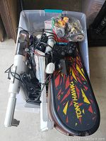 Top view of clear plastic tub containing Tony Hawk skateboard controller, Wii controllers and cables, and other gaming accessories and electronics.